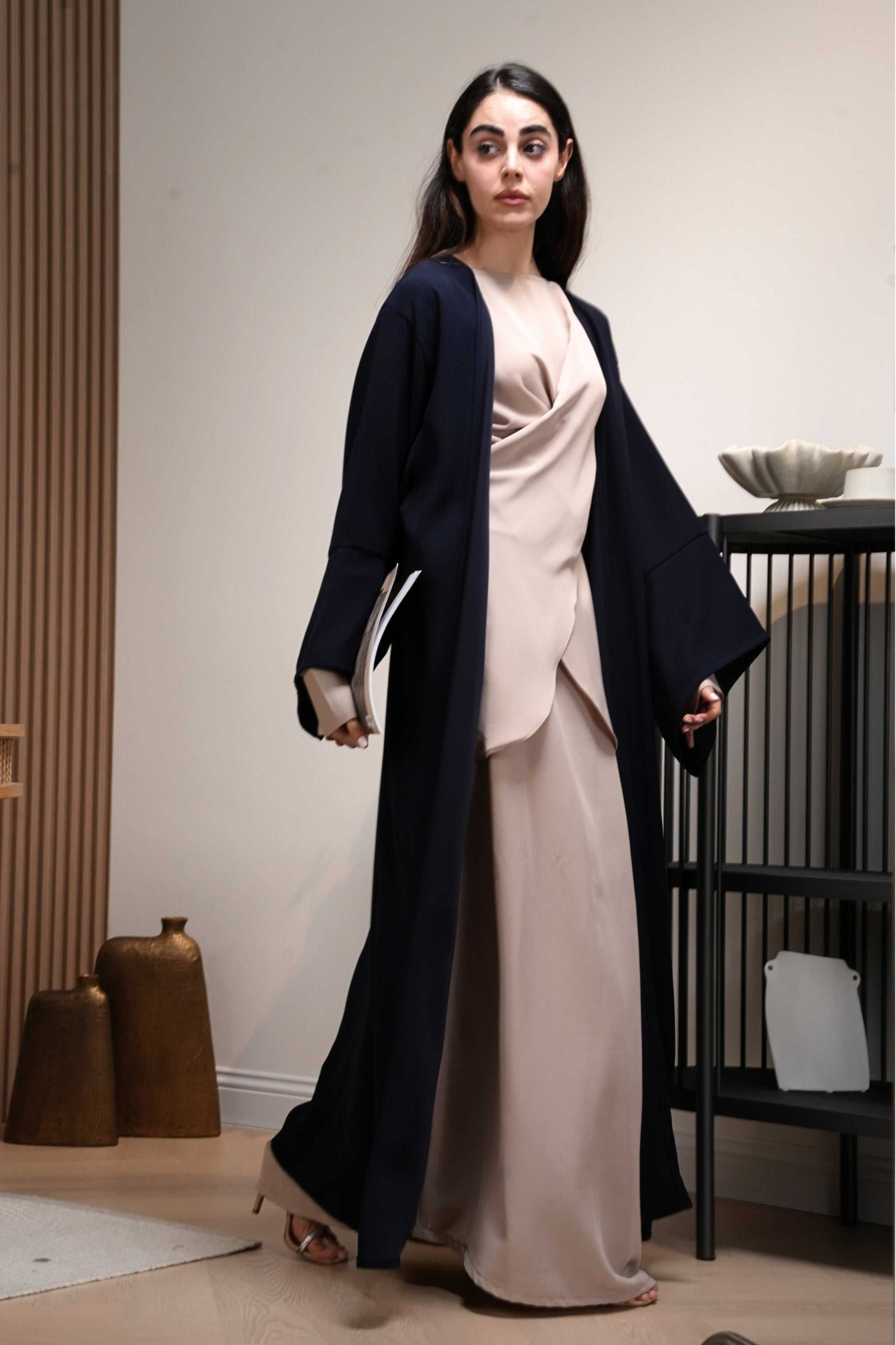 AWAASHI abaya collection, Woman wearing a long navy coat over a beige dress in an indoor setting.